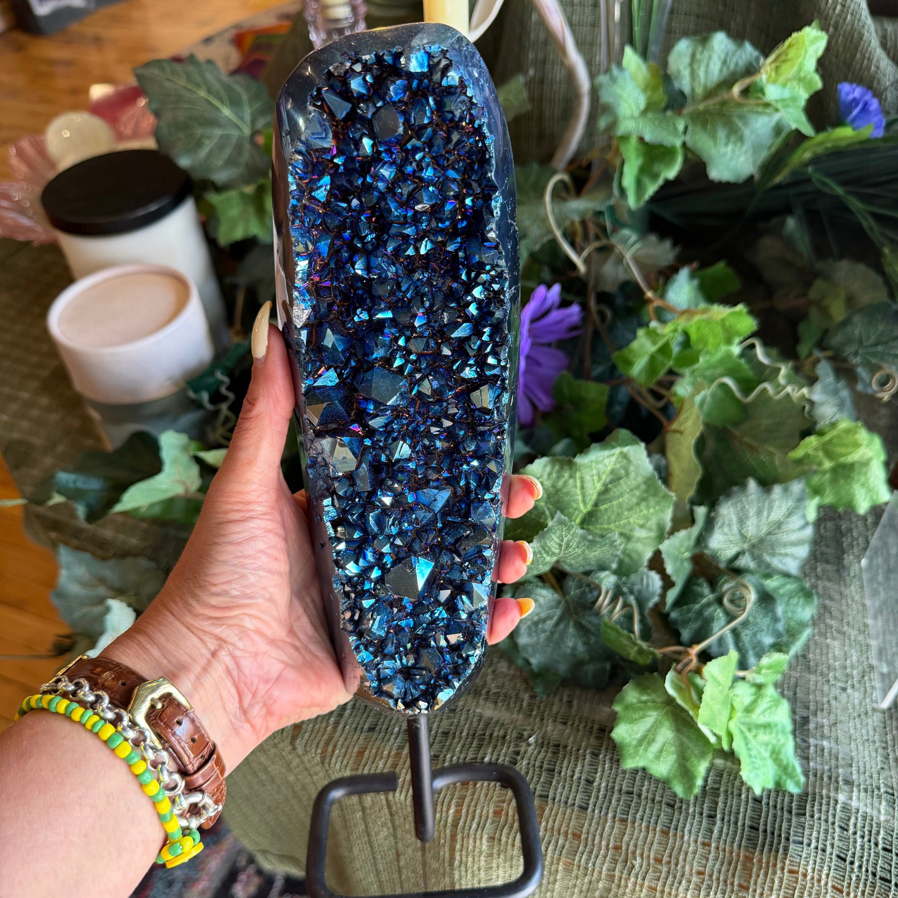 Hand holding a blue crystal on a stand with a candle and plants in the background