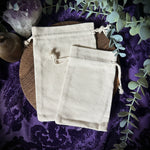Two beige drawstring bags on a purple fabric background with greenery and crystals.