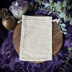 Beige drawstring bag on a wooden surface with purple fabric and green leaves in the background