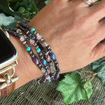 Hand wearing multiple beaded bracelets with a watch and ring, against a natural leafy background