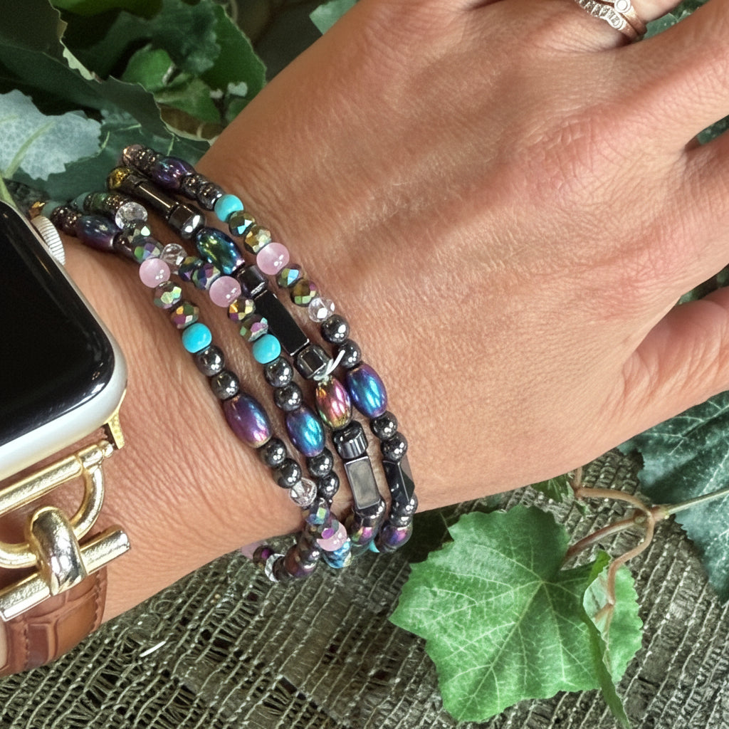 Hand wearing multiple beaded bracelets with a watch and ring, against a natural leafy background