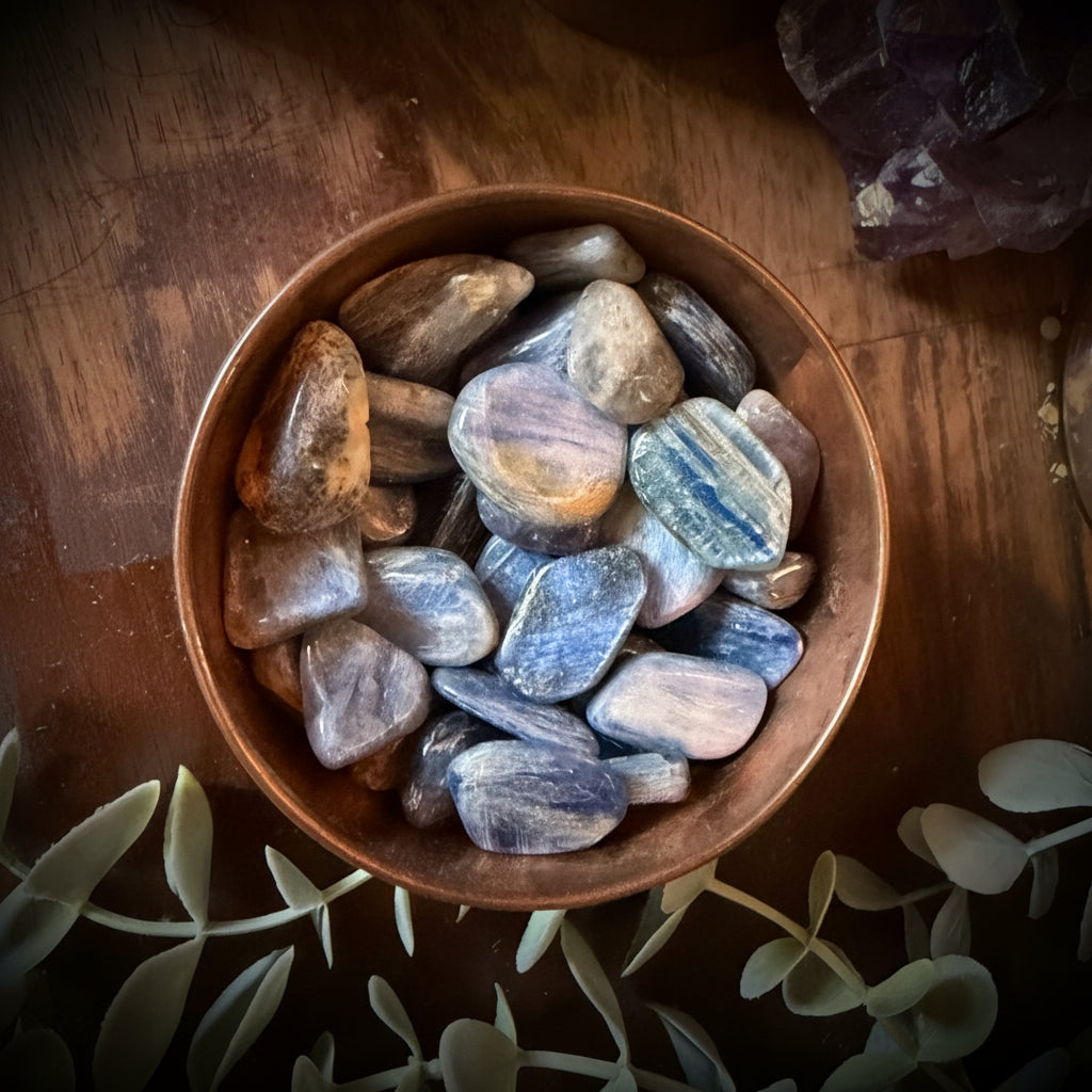 Copper bowl filled with kyanite tumbled stones on a wooden surface with crystals and eucalyptus leaves.