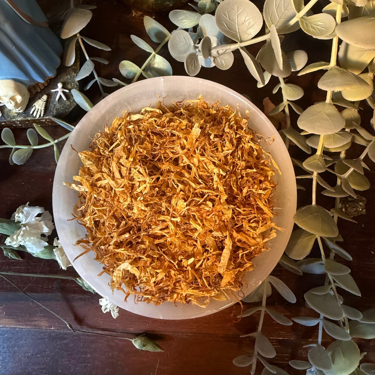 Small white bowl filled with gold-colored calendula flowers on a decorative background with green leaves.