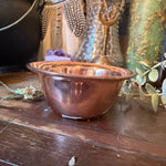 A small copper-colored metal bowl with a ribbed exterior and a flared rim, photographed on a dark wooden surface with decorative objects around it.