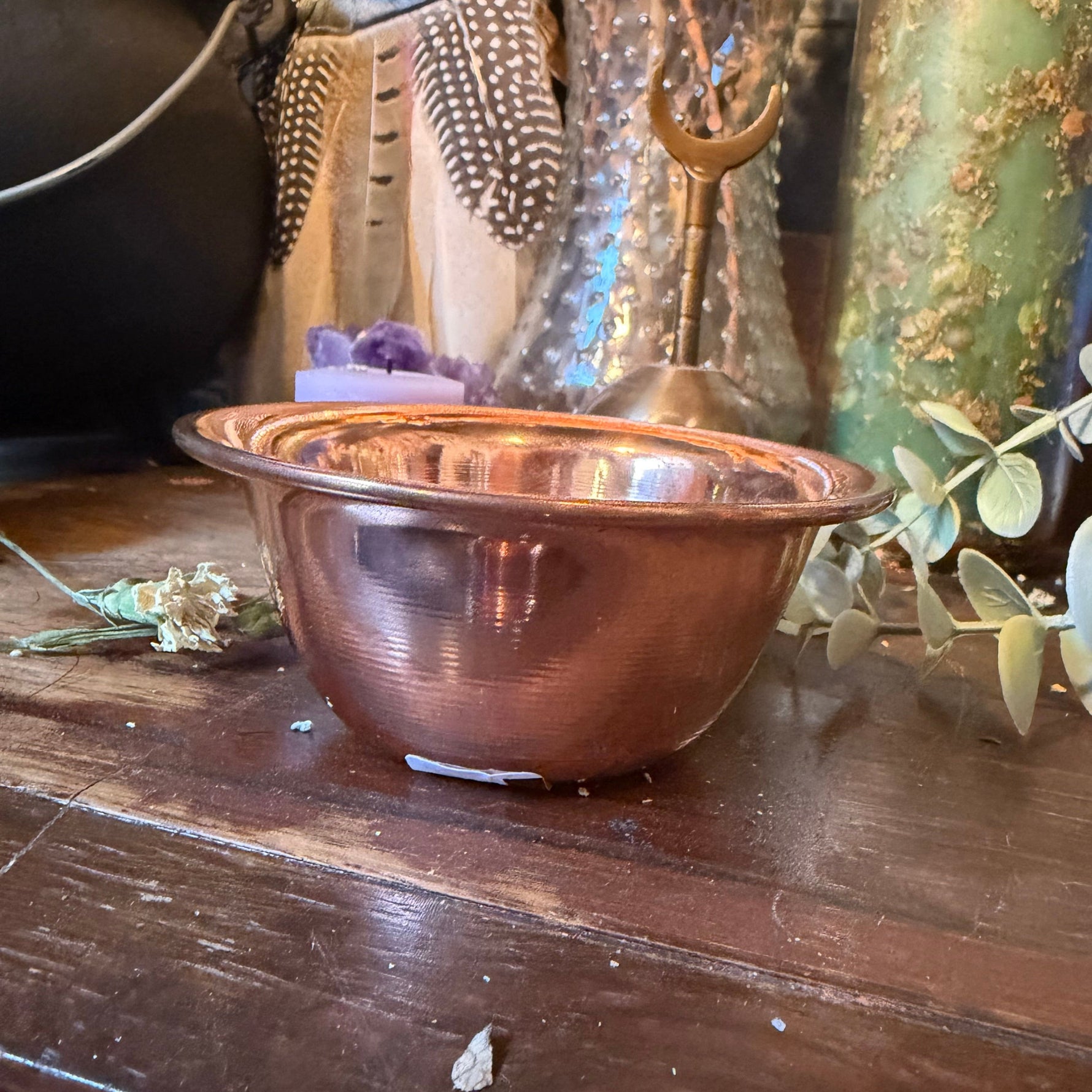 A small copper-colored metal bowl with a ribbed exterior and a flared rim, photographed on a dark wooden surface with decorative objects around it.