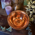 A polished copper bowl with a circular textured design inside, styled on a dark wooden surface with a purple candle, amethyst cluster, brass bell, and green foliage.