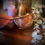 A hand holds a small hammered copper bowl on a rustic wooden shelf, surrounded by decorative items including a purple crystal cluster, a pink candle, a brass bell, and eucalyptus branches.