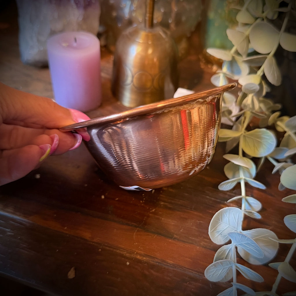 A hand holds a small hammered copper bowl on a rustic wooden shelf, surrounded by decorative items including a purple crystal cluster, a pink candle, a brass bell, and eucalyptus branches.