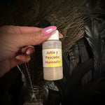 A hand with pink manicured nails holds a small plastic bottle labeled 'Jutia y Pescado Humado' in front of a decorative arrangement of peacock feathers and glass jars.