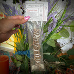 A hand with pink nail polish holds a clear plastic bag containing a bundle of dried muguey root herbs with a white paper header card. The background is a decorative arrangement of purple lavender, white flowers, and green leaves.