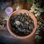 A top-down view of polished black pyrite chunks inside a round terracotta-colored wooden bowl, surrounded by decorative props including a purple crystal cluster, a lavender candle, dried eucalyptus leaves, and a brass object on a rustic wooden surface.