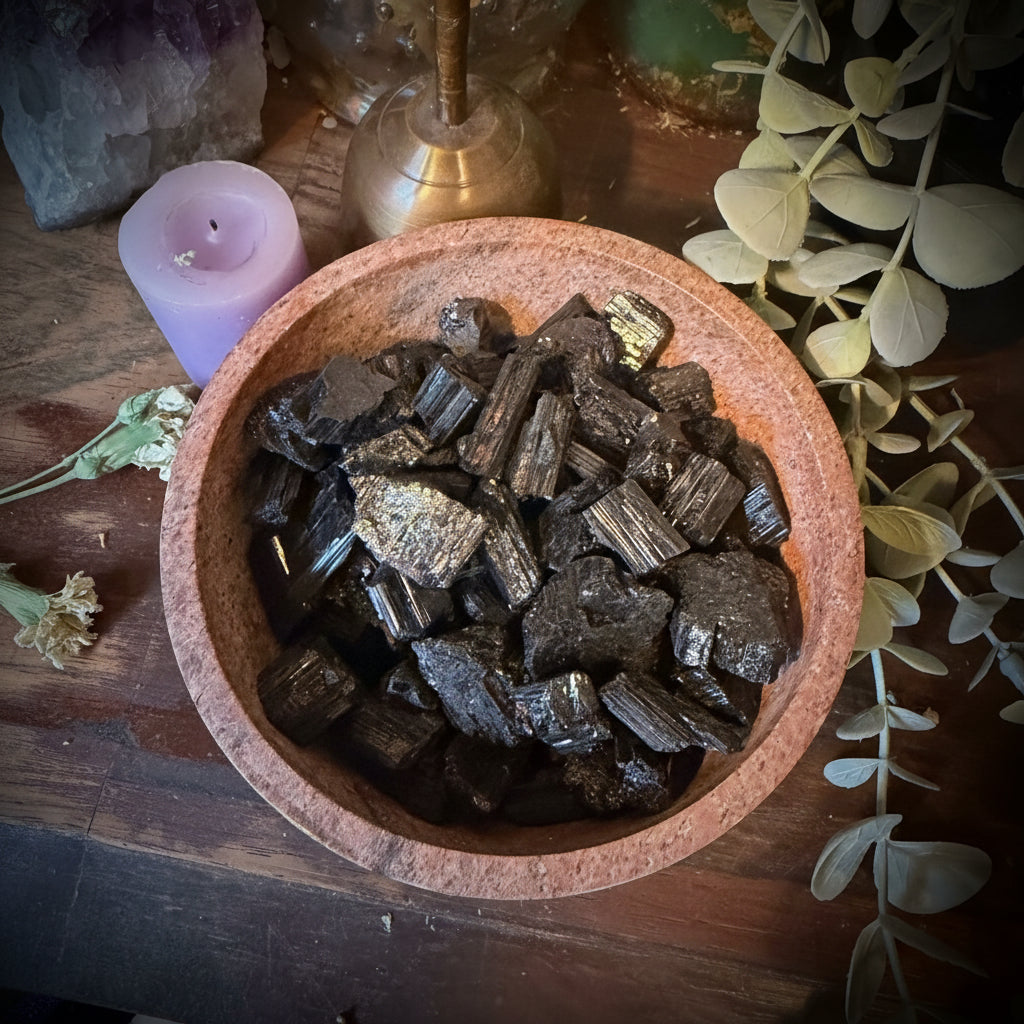 A top-down view of polished black pyrite chunks inside a round terracotta-colored wooden bowl, surrounded by decorative props including a purple crystal cluster, a lavender candle, dried eucalyptus leaves, and a brass object on a rustic wooden surface.