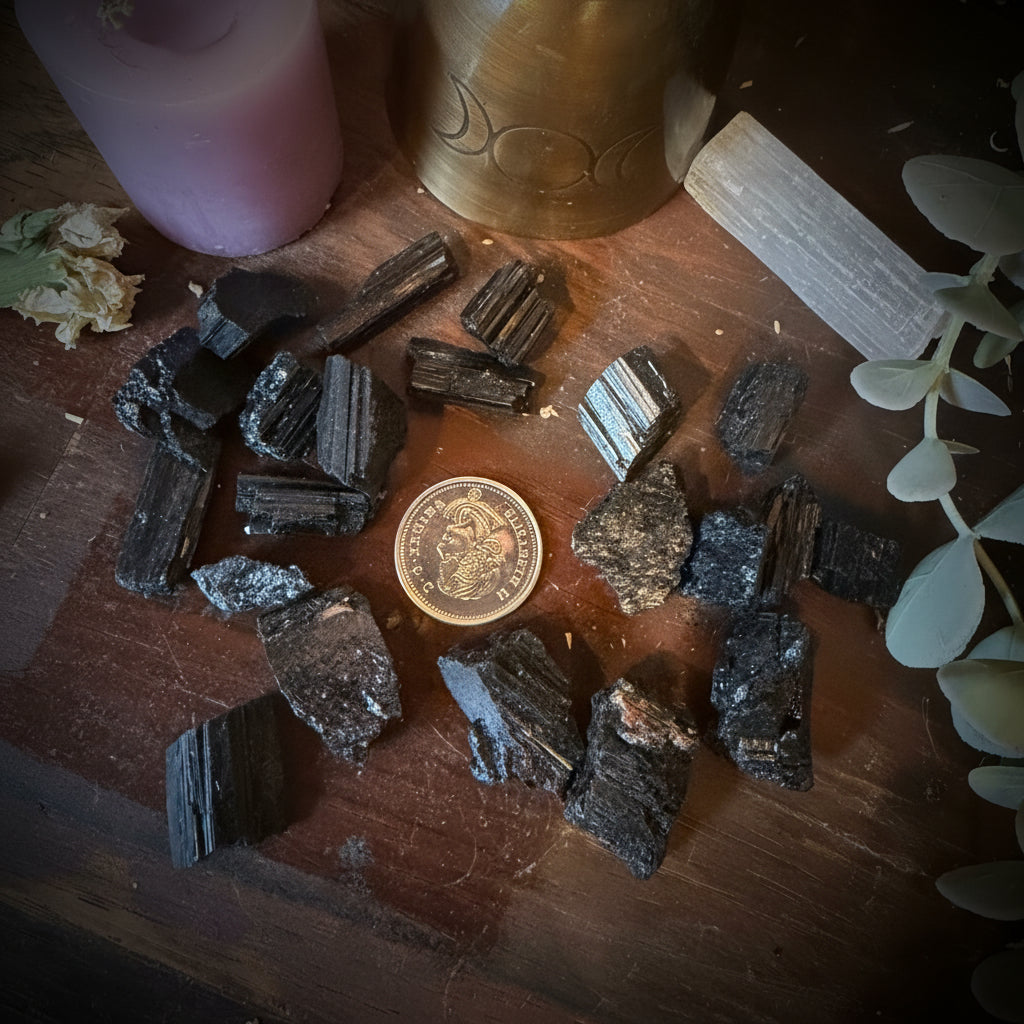 A group of polished black gemstone chips and raw mineral samples arranged on a wooden surface, alongside a pink candle, a brass bottle, a white wood slice, dried flowers, and green leaves.