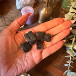 A hand holding several small dark polished crystal chunks, with decorative plants, a purple candle, and crystals on a wooden surface in the background.
