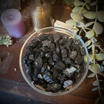 A clear glass bowl filled with dark polished crystal chunks, photographed from above on a wooden surface with decorative dried leaves, a purple candle, and other crystals around it.