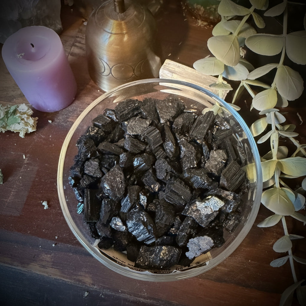 A clear glass bowl filled with dark polished crystal chunks, photographed from above on a wooden surface with decorative dried leaves, a purple candle, and other crystals around it.