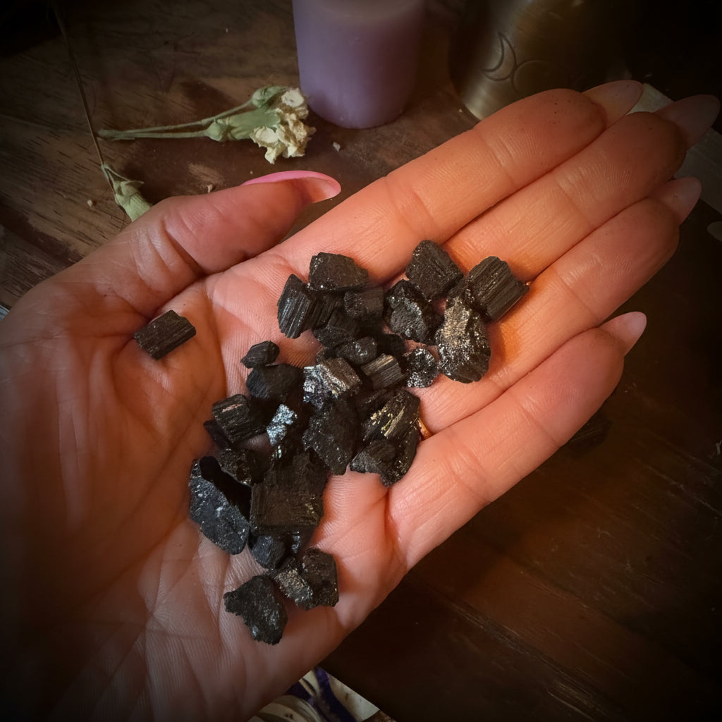 A hand holding small dark polished stone chips over a wooden table, with a lavender candle, crystal cluster, and brass bell in the background.