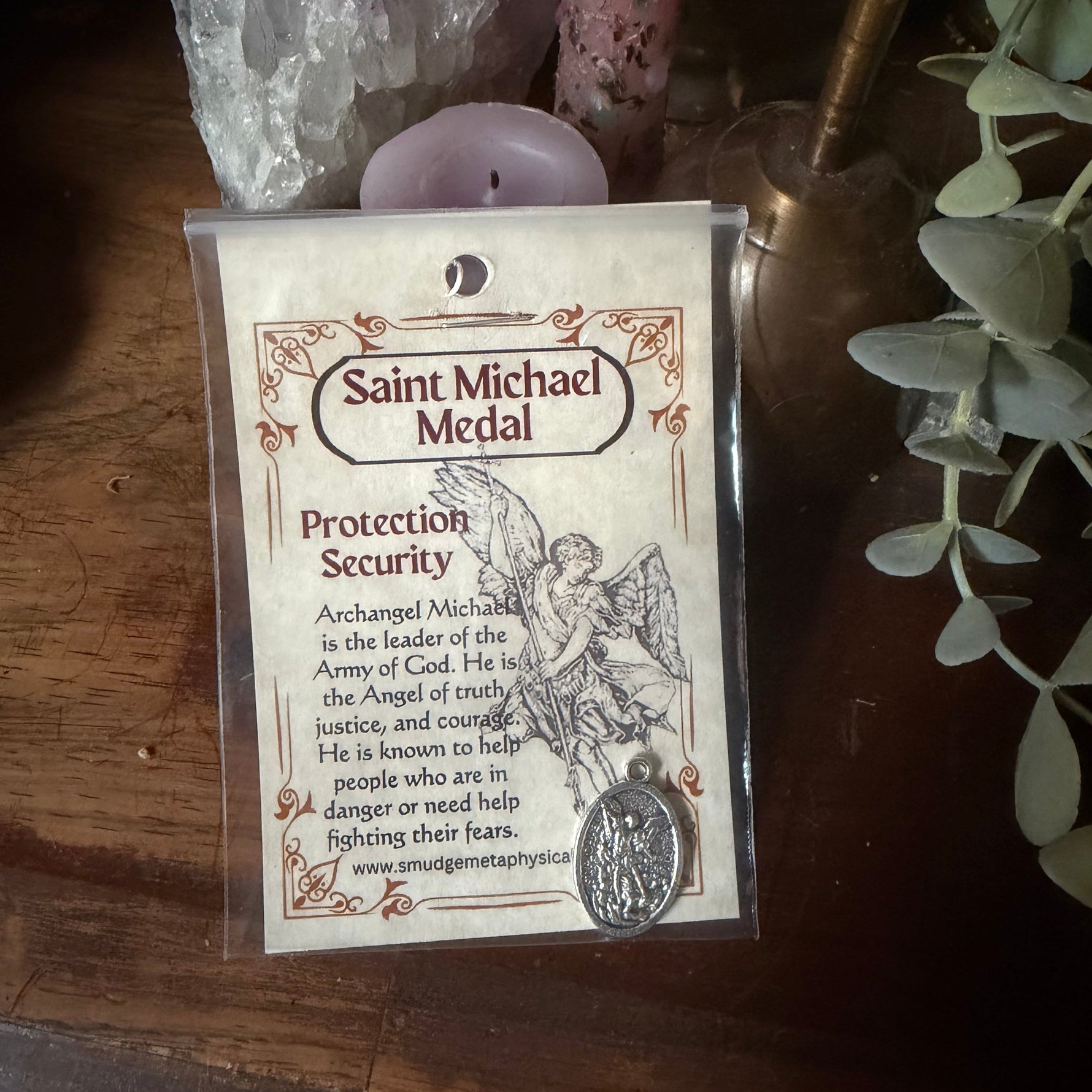 A Saint Michael Medal protection security card in a clear plastic sleeve, placed on a dark wooden surface next to a crystal cluster and a small pink candle.