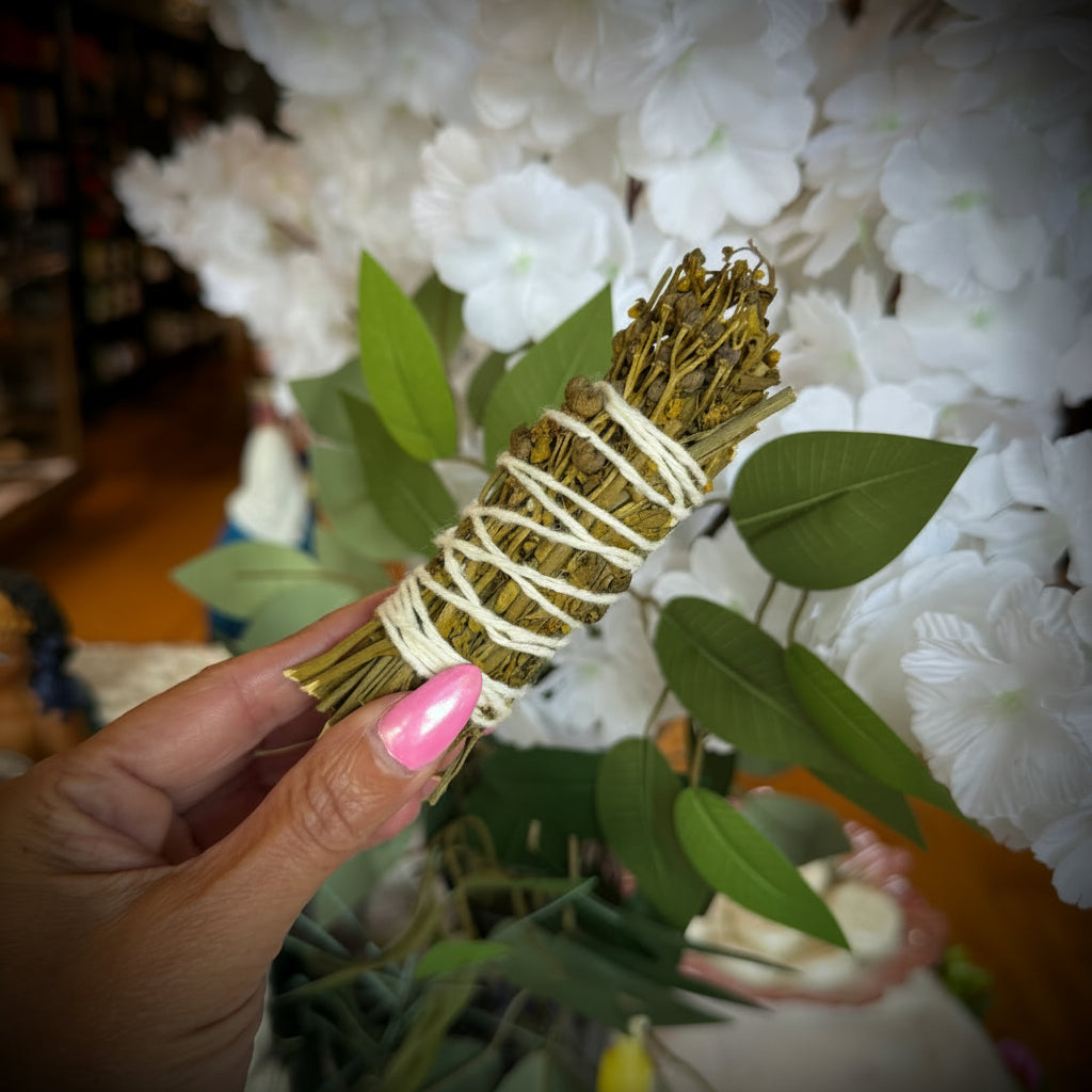 A hand with pink nail polish holds a small sage smudge bundle wrapped in white string, with white flowers and green leaves in the background.