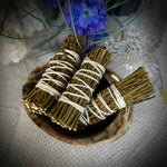 Two dried sage smudge bundles wrapped in white string, resting in a small natural shell dish on a textured white surface, with blue flowers and glass decor in the background.