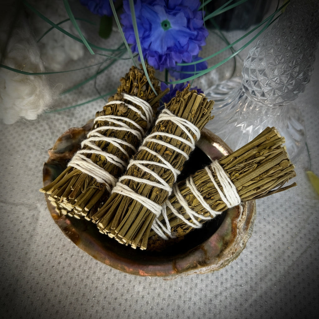 Two dried sage smudge bundles wrapped in white string, resting in a small natural shell dish on a textured white surface, with blue flowers and glass decor in the background.