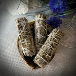Three dried sage smudge sticks wrapped in white string, arranged in a small bowl on a textured white surface, with decorative blue flowers and a white textured background.