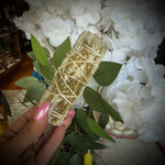 A hand with pink manicured nails holds a sage smudge bundle wrapped in string, positioned in front of a large arrangement of white flowers and green leaves.