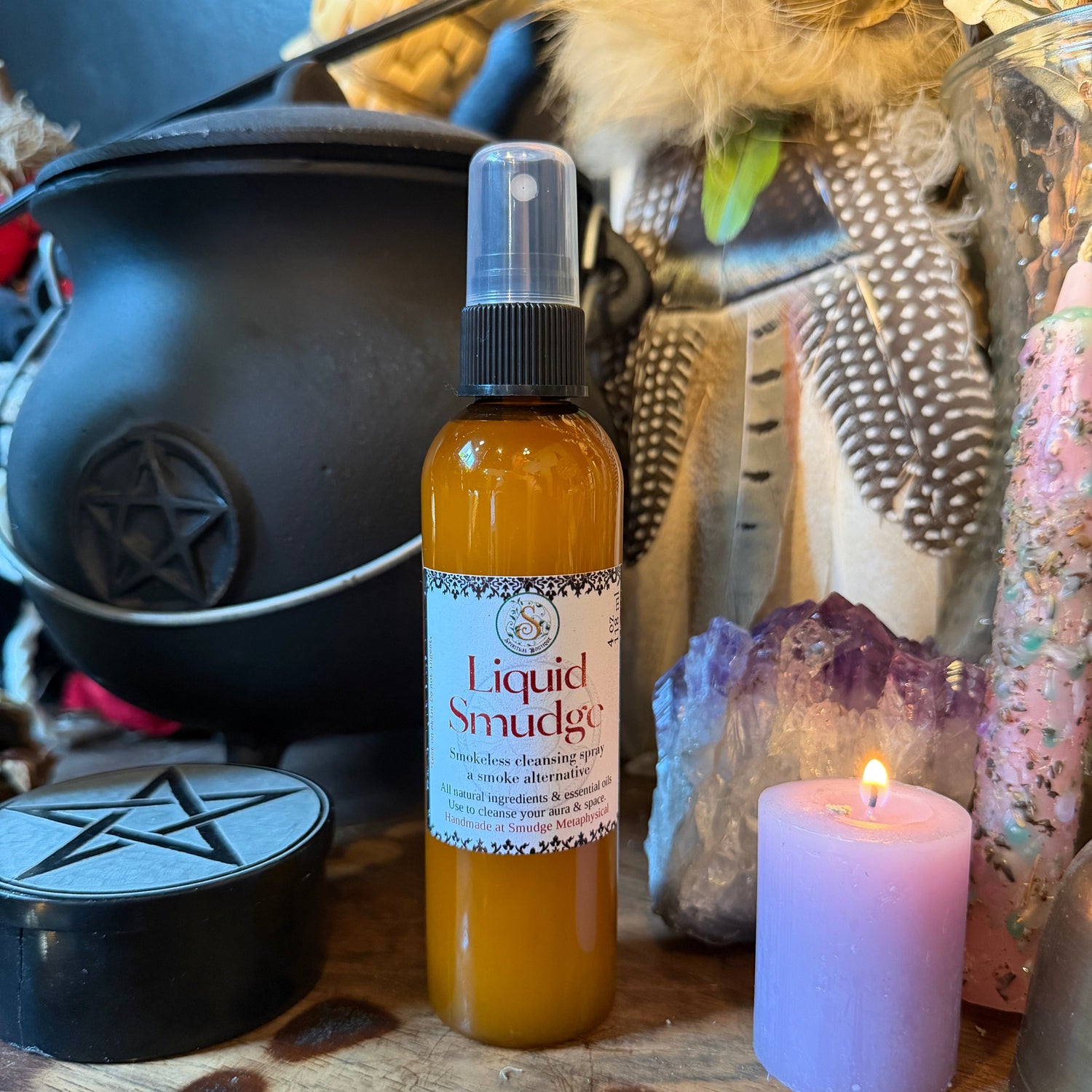 A bottle of Liquid Smudge room spray, with amber liquid and a white label, styled on a wooden surface with a black cauldron, purple crystal, and lit lavender candle.