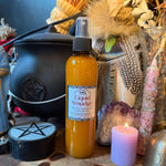 A bottle of 'Sacred Liquid Smudge' essential oil spray with amber-colored liquid inside, displayed on a wooden surface surrounded by mystical props including a black cauldron, a purple candle, dried flowers, and a pentacle cup.