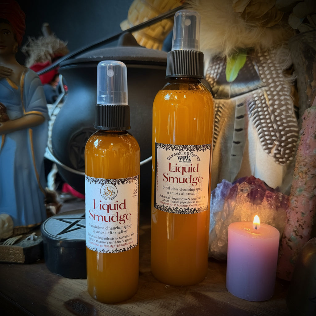 Two amber glass spray bottles labeled 'Liquid Smudge' with white labels and black spray tops, displayed on a wooden surface with dried flowers, feathers, crystals, and a pink candle in the background.