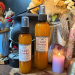 Two amber glass spray bottles labeled 'Liquid Smudge' with white labels and black spray tops, displayed on a wooden surface with dried flowers, feathers, crystals, and a pink candle in the background.