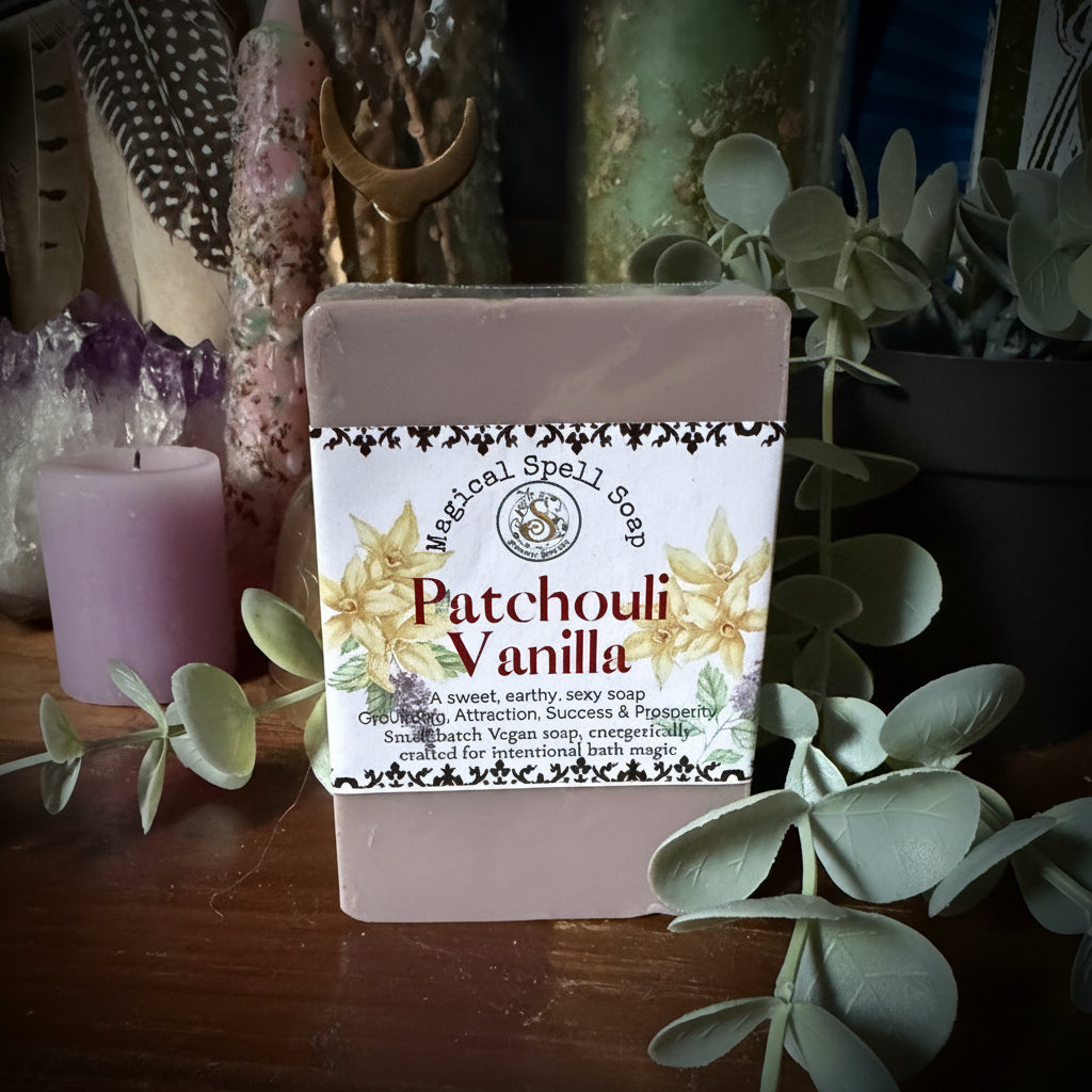 A rectangular bar of soap labeled 'Magic Soap Patchouli Vanilla' resting on a dark wooden surface, surrounded by green eucalyptus leaves, crystals, and decorative items.