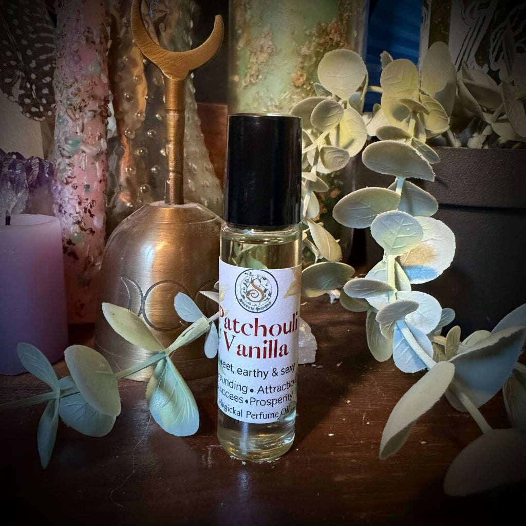 A small glass roller bottle labeled 'witchboul vanilla' with a black cap, placed on a wooden surface surrounded by dried eucalyptus leaves, crystals, and decorative objects.