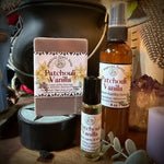 A bath and body gift set from Sweet Wildflower Soap featuring a Patchouli Vanilla soap bar, a matching spray bottle, and a roller bottle, arranged on a rustic wooden surface with a black cauldron and dried botanicals.