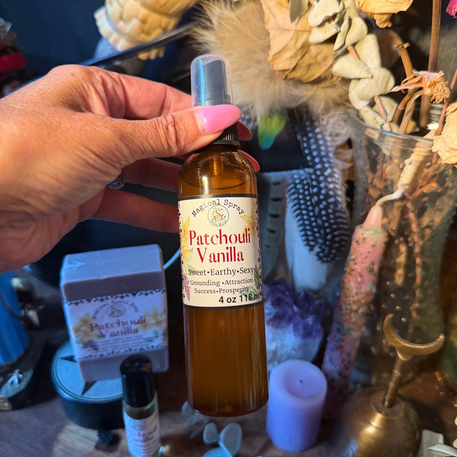 A hand with pink nail polish holds a bottle of Patchouli Vanilla essential oil against a rustic, bohemian-themed background with dried flowers, crystals, and candles.