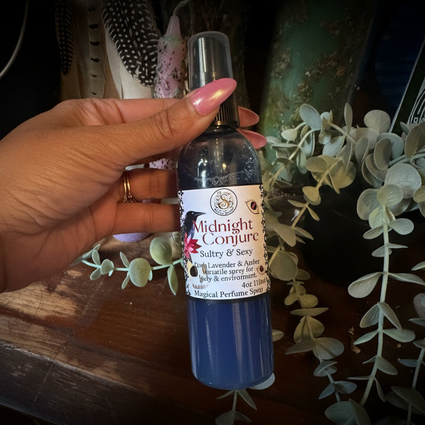 A hand holding a bottle of Midnight Conjure Body & Spray, showing the blue cap and white label, set against a rustic wooden background with eucalyptus leaves.