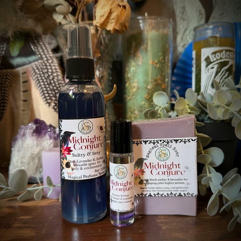 A Midnight Conjure salve and soap set displayed on a wooden surface, featuring a blue spray bottle, a small lavender soap vial, and a pink soap bar with branded labels, surrounded by dried flowers and botanical decor.