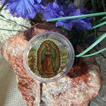 A round clear plastic jar containing a colorful religious medallion or prayer card featuring Our Lady of Guadalupe, styled on a textured surface with purple flowers and a polished stone.