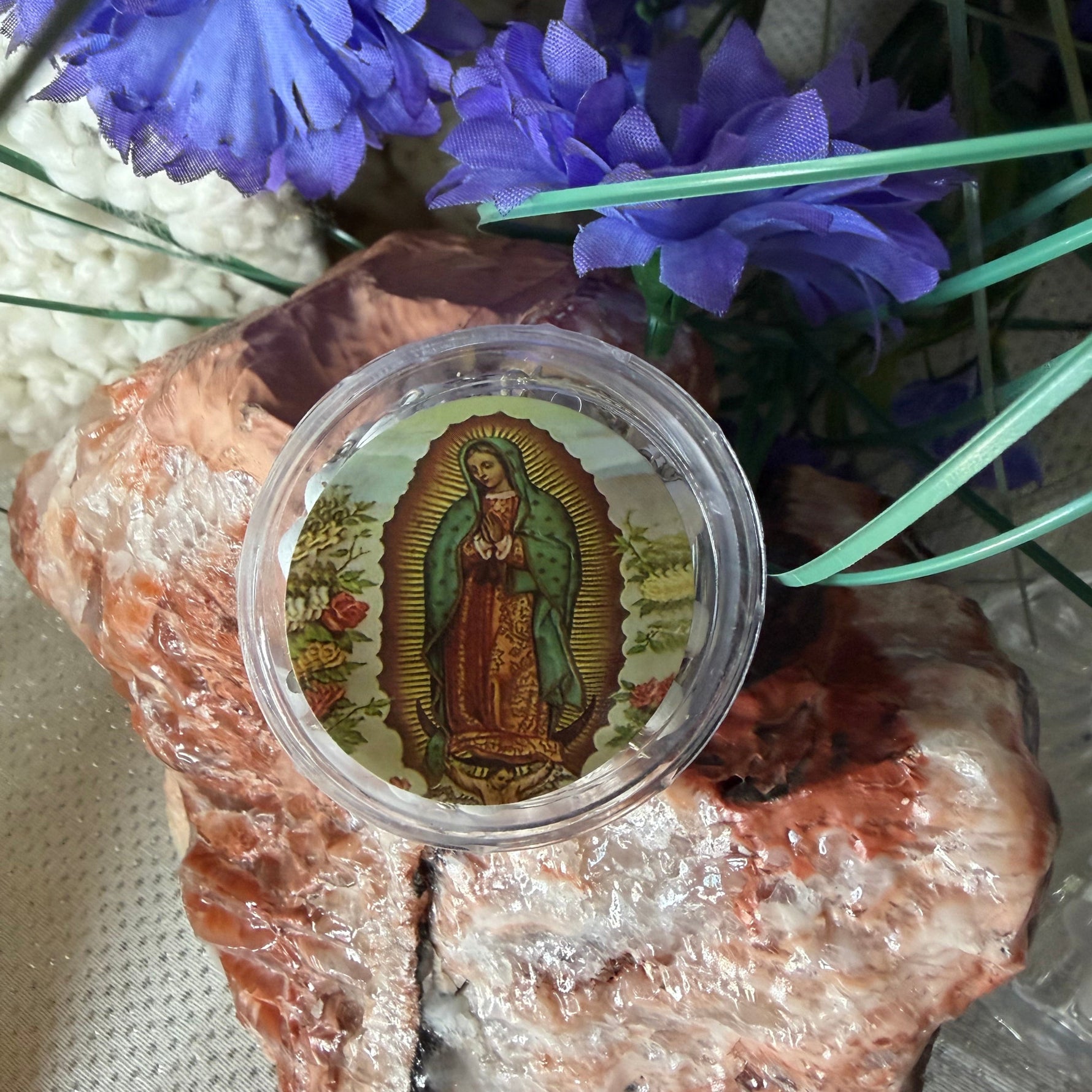 A round clear plastic jar containing a colorful religious medallion or prayer card featuring Our Lady of Guadalupe, styled on a textured surface with purple flowers and a polished stone.