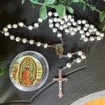 A white beaded rosary with a silver-tone crucifix and a small round medallion featuring Our Lady of Guadalupe, arranged on a dark tabletop with green leaves around it.