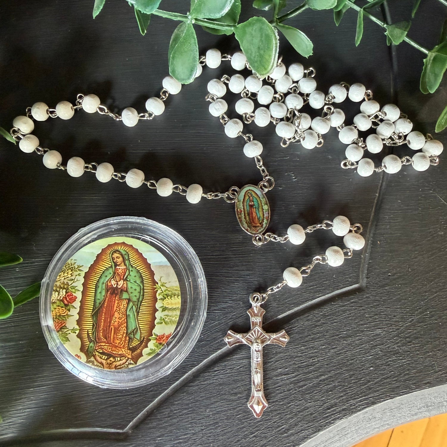 A white beaded rosary with a silver-tone crucifix and a small round medallion featuring Our Lady of Guadalupe, arranged on a dark tabletop with green leaves around it.