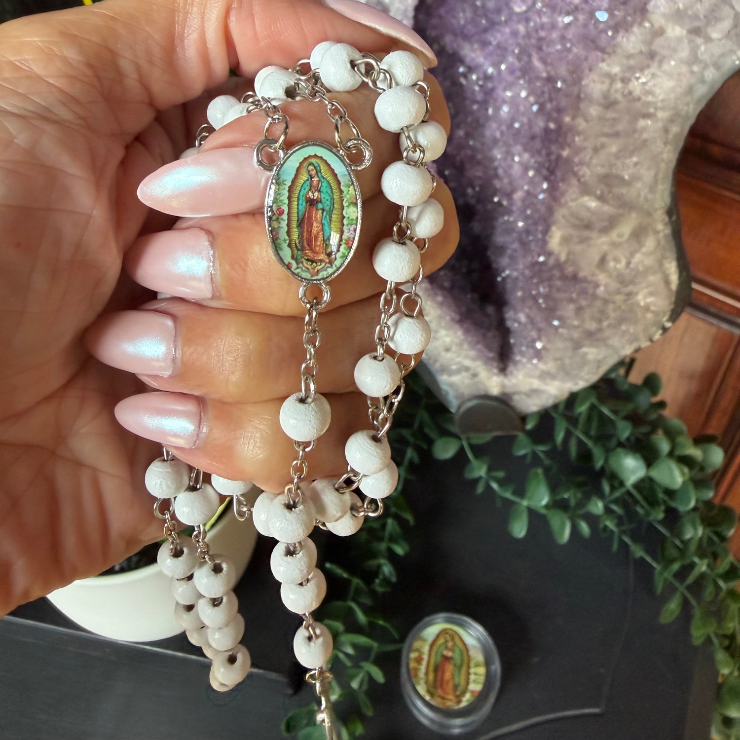 A hand with long pink manicured nails holds a white beaded rosary with a colorful Our Lady of Guadalupe medal. The background features a large purple amethyst geode, green foliage, and a dark surface.