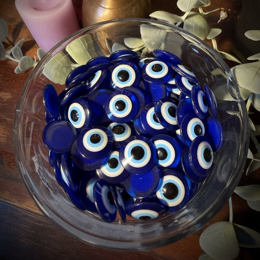 A clear glass bowl filled with round blue glass evil eye charms, photographed from above on a wooden surface with decorative leaves, crystals, and a pink candle nearby.