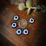 Five blue and white glass evil eye beads arranged around a US quarter coin on a dark wooden tabletop, with decorative eucalyptus leaves and a pink candle partially visible.