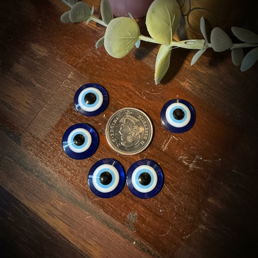 Five blue and white glass evil eye beads arranged around a US quarter coin on a dark wooden tabletop, with decorative eucalyptus leaves and a pink candle partially visible.