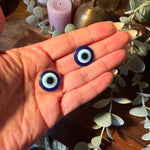 Two blue and white evil eye beads held in a person's open palm, with decorative eucalyptus leaves and candles in the background.