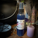 A bottle of Midnight Conjure body spray with dark blue liquid and a white label, displayed on a wooden surface with a black cauldron, a lavender candle, dried feathers, and other mystical props.