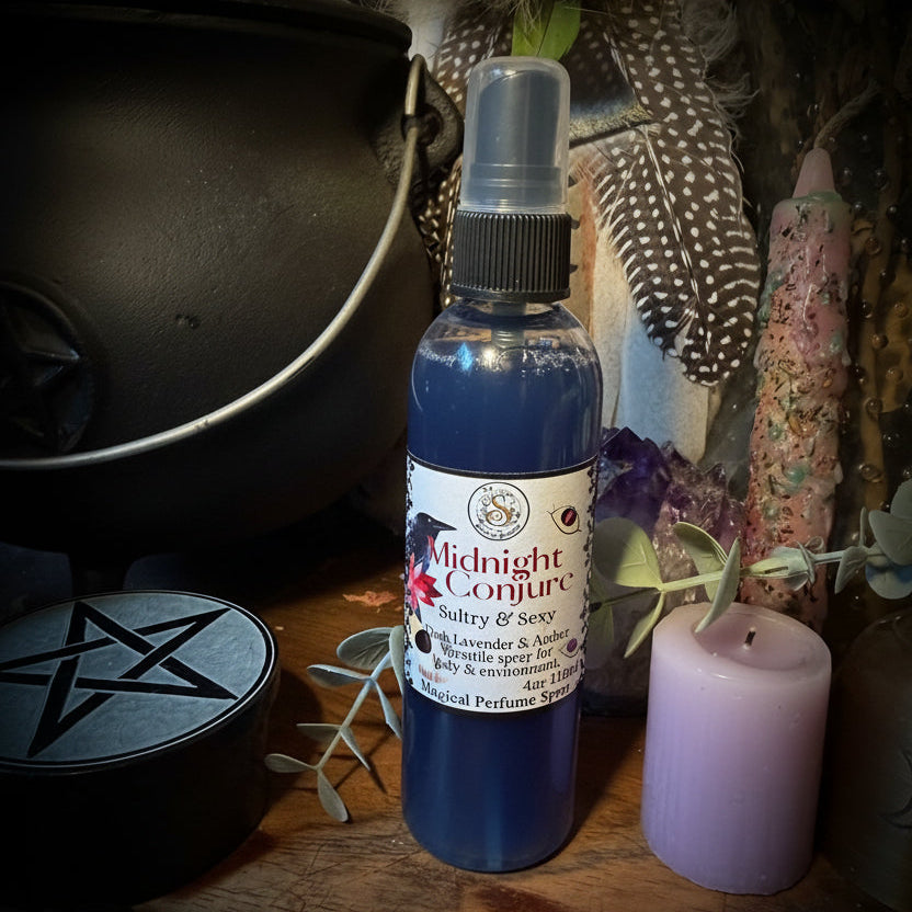 A bottle of Midnight Conjure body spray with dark blue liquid and a white label, displayed on a wooden surface with a black cauldron, a lavender candle, dried feathers, and other mystical props.