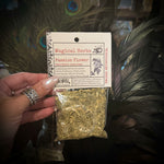 A hand holding a small clear plastic bag of dried 'Passion Flower' herbs with a white paper header card labeled 'Magical Herbs'. The background is a decorative display with peacock feathers and glass jars.