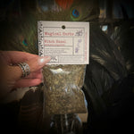 A hand holding a clear plastic bag of dried witch hazel herbs with a white paper label that reads 'Magical Herbs' and 'Witch Hazel'. The background features decorative black feathers and glass jars containing dried botanicals.
